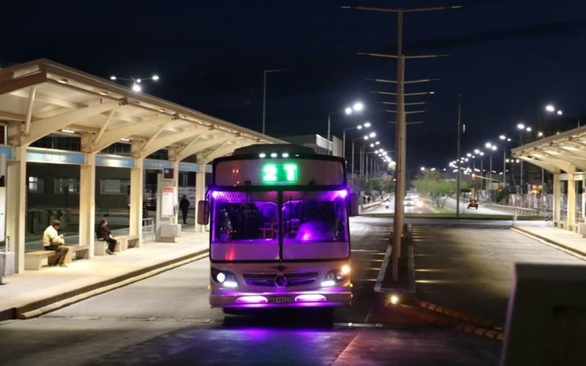 Un colectivo de la línea 21 con luces encendidas llega de noche a una parada techada en San Juan.