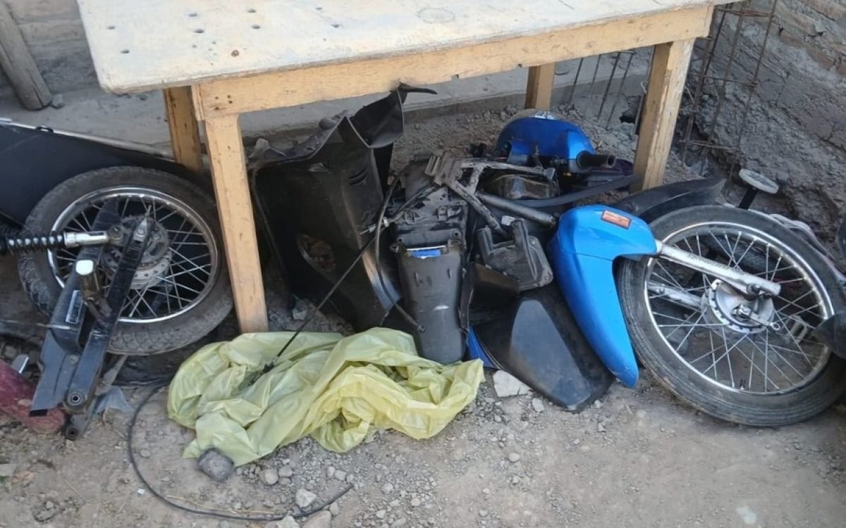 Partes de una moto azul y negra desarmada, con ruedas, motor y plásticos, en el fondo de una vivienda.