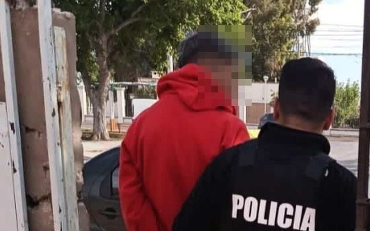 Un policía de espaldas acompaña a un hombre con buzo rojo, con el rostro pixelado, junto a un portón.
