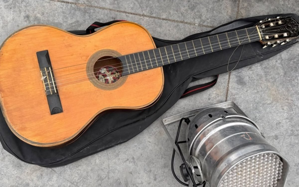 Una guitarra criolla sobre su funda negra y una lámpara LED plateada, apoyadas en el piso.