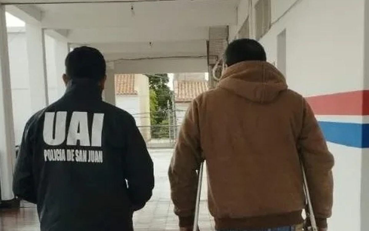 Un efectivo de la UAI de la Policía de San Juan camina junto a un hombre por un pasillo, de espaldas.